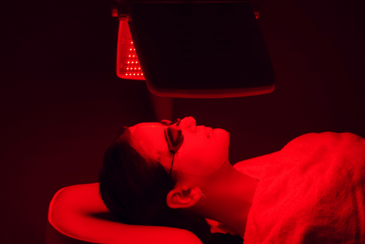 Woman in her 40s or 50s lying under red light therapy, wearing eye protection in a calm, softly lit setting.